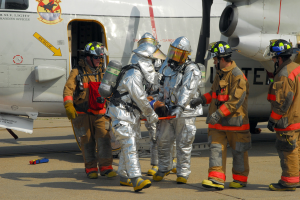 Eine Gruppe von Feuerwehrleuten in Helmen und Schutzausrüstung steht vor einem Flugzeug auf der Rollbahn, wobei einer ein Objekt hält und verschiedene Gegenstände um sie herum auf dem Boden verstreut sind.