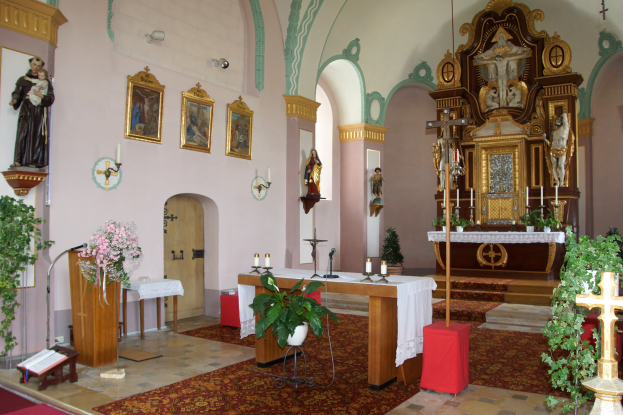 Das Innere einer Kirche mit einem Kreuz auf dem Altar, umgeben von Zimmerpflanzen, Blumensträußen, Kerzen mit Ständern, Tischen, Stühlen, Teppichen auf dem Boden, Statuen, Fotorahmen an der Wand, einer Tür und anderen Gegenständen.