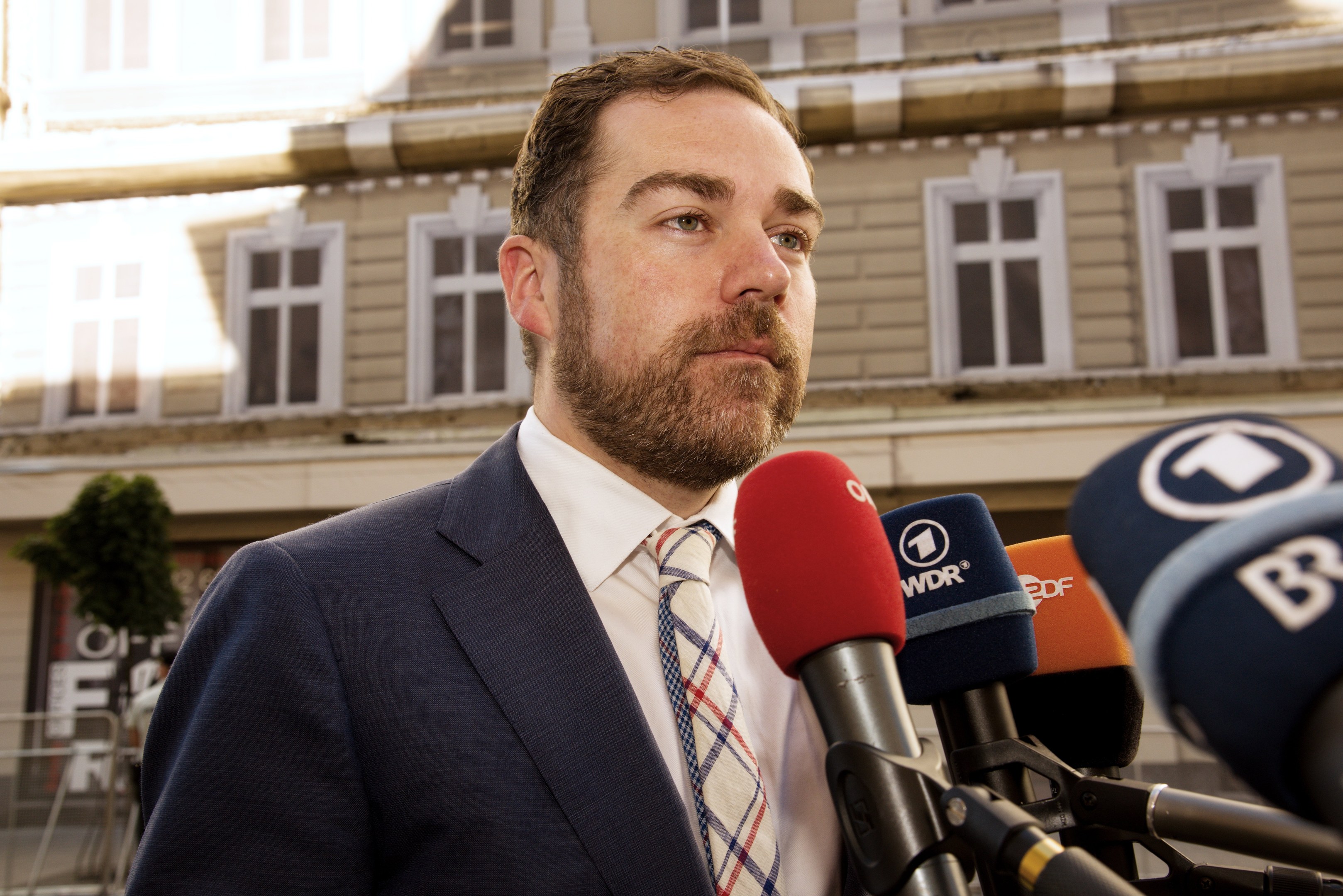 Ein Mann im Anzug spricht zu Medien vor einem Gebäude, wahrscheinlich im Zusammenhang mit einer dreijährigen Haftstrafe.