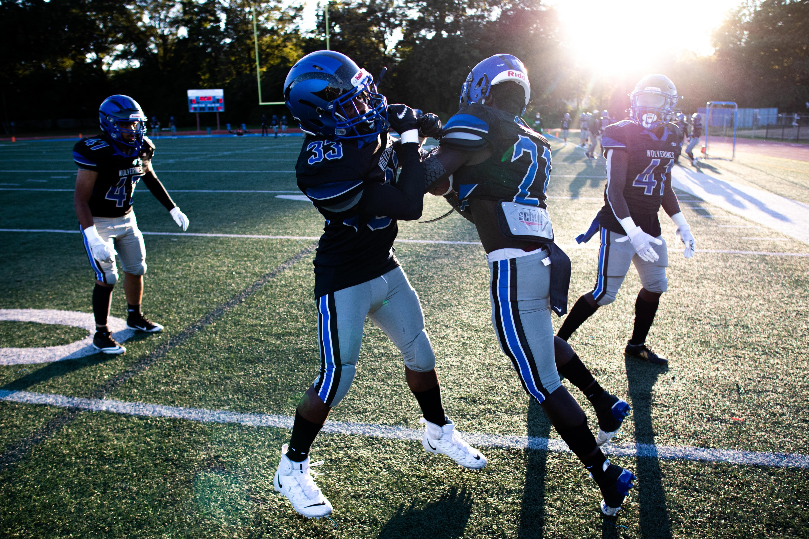 Gruppe junger Männer in Helmen und Schuhen, die auf einem von Bäumen umgebenen Feld unter einem klaren blauen Himmel Fußball spielen.