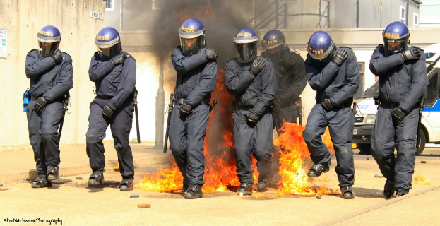 Menschen in Helmen vor einem Feuer mit Gegenständen auf dem Boden, Gebäuden im Hintergrund, einem Fahrzeug, einem Plakat und einer Tafel an der linken Wand und Text unten.
