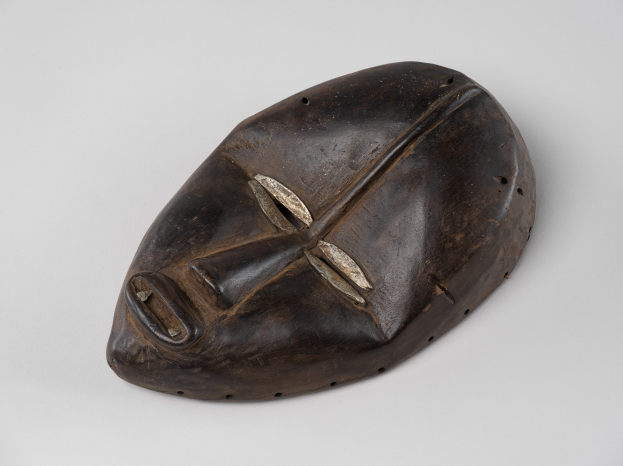 Eine braune hölzerne Maske mit einem geschnitzten Gesicht und detaillierten Verzierungen liegt auf einer weißen Oberfläche.