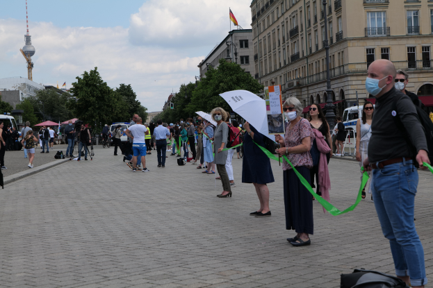 Eine Gruppe von Menschen marschiert auf einer Straße in Berlin, Deutschland, mit Schildern und Transparenten, einige tragen Masken und tragen Taschen oder Schirme, mit Bäumen, Gebäuden, geparkten Fahrzeugen und einem flaggentoppten Turm im Hintergrund unter einem bewölkten Himmel.