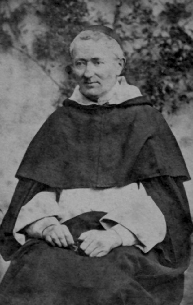 Altes Schwarz-Weiß-Foto eines Mannes in Priesterrobe, der vor Bäumen sitzt, in schwarzer Robe mit weißem Kragen und Manschetten, die Hände zum Gebet gefaltet, von warmem Licht beleuchtet mit friedlichem Gesichtsausdruck.