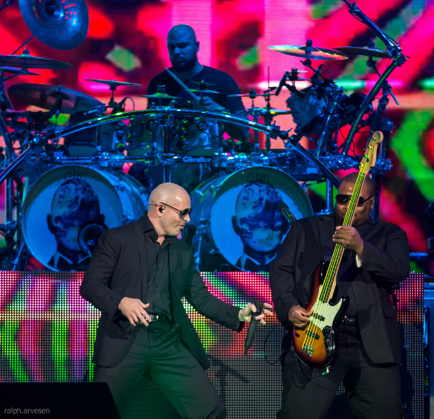 Ein Mann in der rechten Ecke spielt eine Gitarre, ein anderer Mann in einem schwarzen Blazer hält ein Mikrofon und lächelt, während ein Mann in einem schwarzen T-Shirt Schlagzeug spielt, alles vor einem bunten Hintergrund auf einem Musikkonzert.