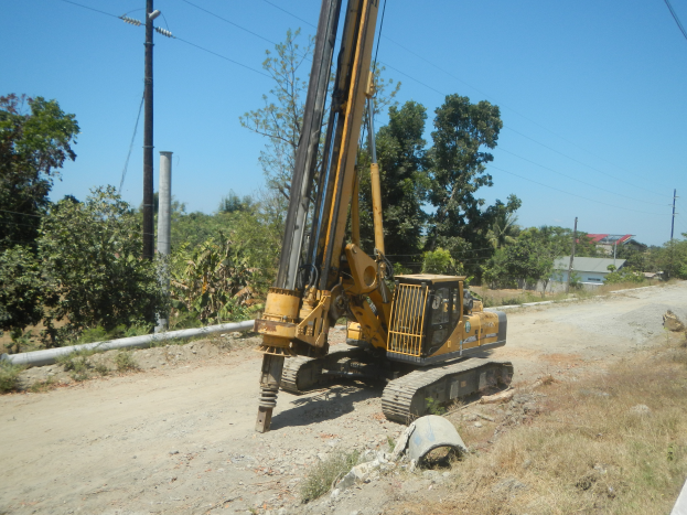 Eine Bohranlage ist auf der Seite einer Schotterstraße positioniert und wird von Gras, Steinen, einem Rohr, einer Gruppe von Bäumen, Strommasten mit Drähten, einem Haus mit Dach und einem bewölkten Himmel umgeben.