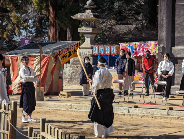 Eine Gruppe von Menschen in traditioneller Kleidung steht im Freien in Kyoto, einige tragen Masken und halten Holzstöcke, während Stühle, Banner und ein Zelt gegen einen klaren blauen Himmel zu sehen sind.