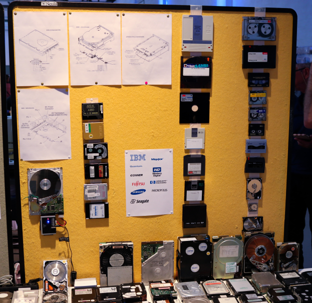Ein Tisch voller verschiedener Festplatten, eine Person rechts daneben mit einem Gegenstand in der Hand und eine Tafel mit Papieren im Hintergrund.