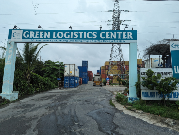 Einfahrt zum Green Logistics Center in Ho-Chi-Minh-Stadt, Vietnam, mit einem Torbogen mit Text, flankiert von Grünpflanzen, mit Containern, einem Fahrzeug und einem Turm mit Drähten im Hintergrund unter einem sichtbaren Himmel.