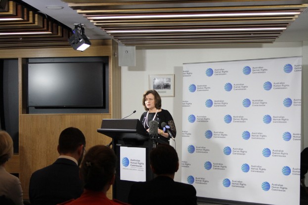 Eine Frau steht auf einer Bühne und spricht in ein Mikrofon, vor ihr sitzt ein Publikum, hinter ihr eine Wand mit einem Banner und ein Fernseher auf einem Holzwand, sowie Lampen an der Decke.