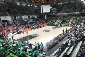 Große Halle mit Zuschauern bei einem Basketballspiel, mit sichtbarem Basketballkorb, Sitzbereichen und Deckenbeleuchtung und -bildschirm.