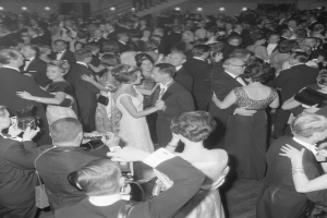 Ein Schwarz-Weiß-Foto von einer Gruppe von Menschen, die auf einer Tanzfläche tanzen, einige halten Kameras in der Hand, mit Treppen und Lichtern im Hintergrund.