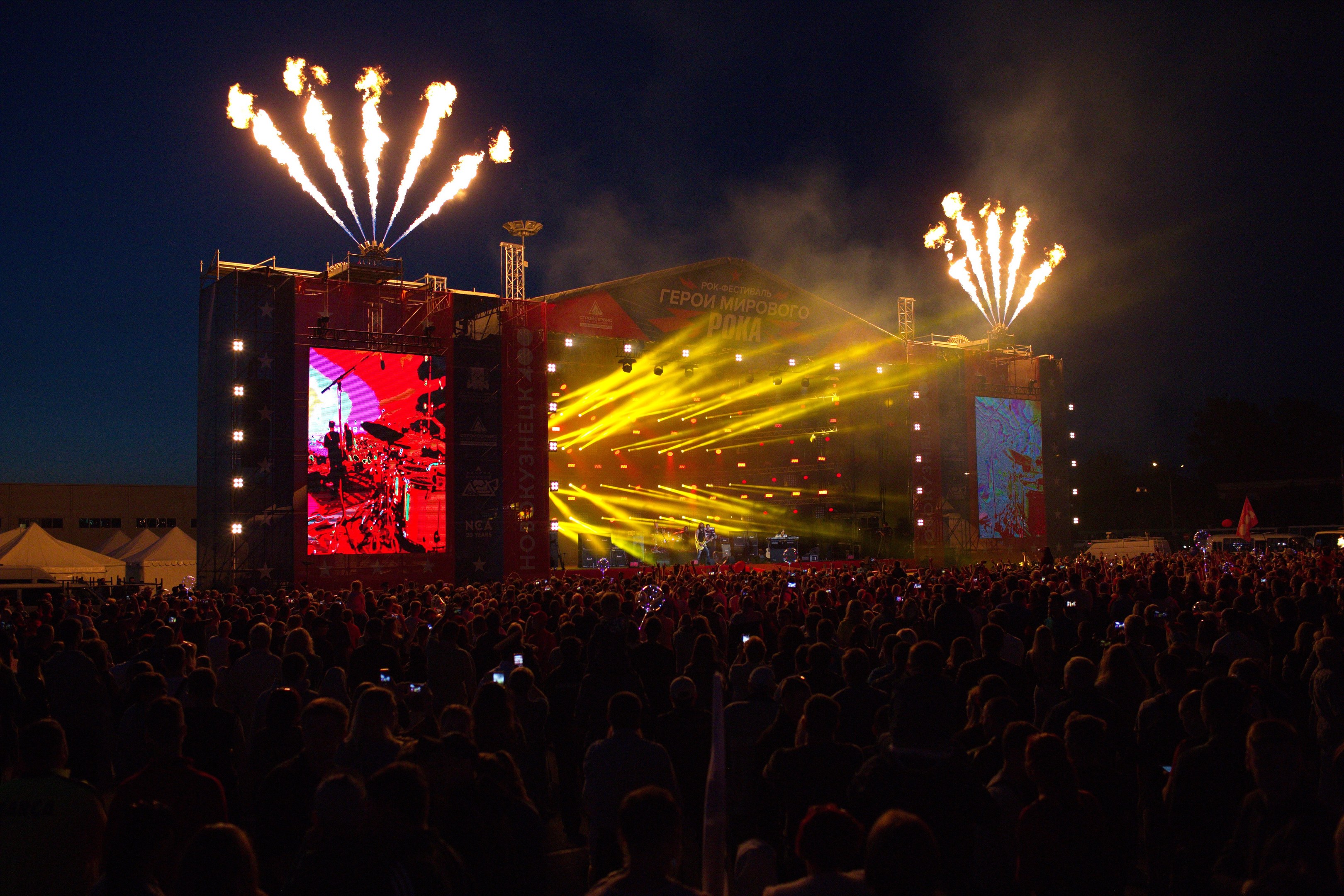 Eine große Menge bei einem Musikfestival schaut auf einer hell beleuchteten Bühne mit Bannern, umgeben von Zelten und Feuerwerk am Nachthimmel.