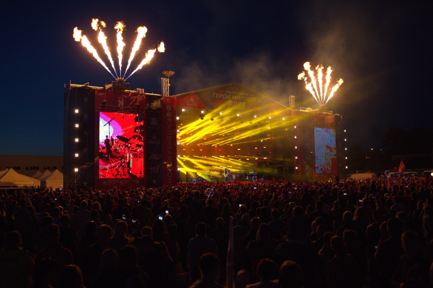 Eine große Menge bei einem Musikfestival schaut auf einer hell beleuchteten Bühne mit Bannern, umgeben von Zelten und Feuerwerk am Nachthimmel.
