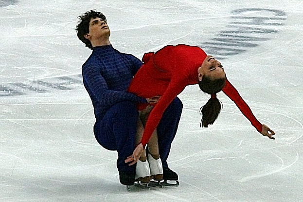 Ein Mann in einem blauen Kleid und eine Frau in einem roten Kleid tanzen gemeinsam auf einem Eisstadion, ihre Arme und Beine ineinander verschlungen, perfekt synchronisiert.