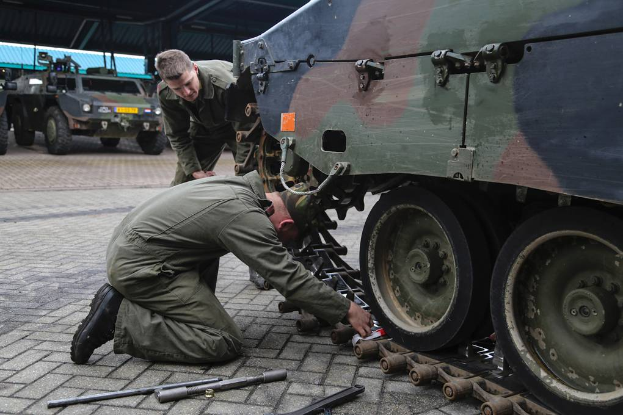 Zwei Männer in Militäruniform arbeiten an einem Fahrzeug in einer Garage, mit Werkzeugen, die auf dem Boden verstreut sind, und anderen Fahrzeugen und einer Hütte im Hintergrund.