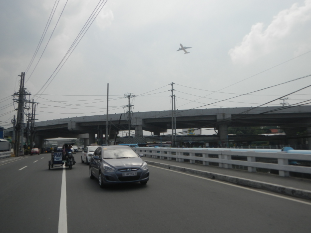 Ein Auto fährt auf einer Autobahn mit einer Brücke im Hintergrund, flankiert von Strommasten mit Überlandleitungen, während ein Flugzeug am Himmel darüber fliegt.