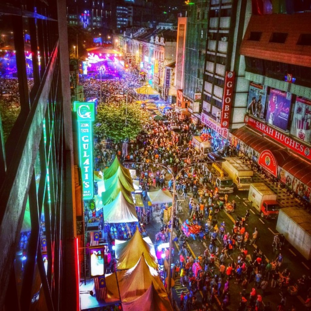 Eine lebhafte Karnevalsszene mit einer großen Menschenmenge auf der Straße, umgeben von Zelten, Geschäften, Gebäuden und Bannern, mit einer hell erleuchteten Fläche im Hintergrund.