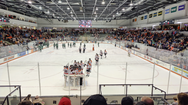 Ein Hockey-Spiel in einer Eishalle mit Spielern, Tor, Zuschauern in der Tribüne, Hallenbeleuchtung, Großbildschirm und Bannern an der Decke.
