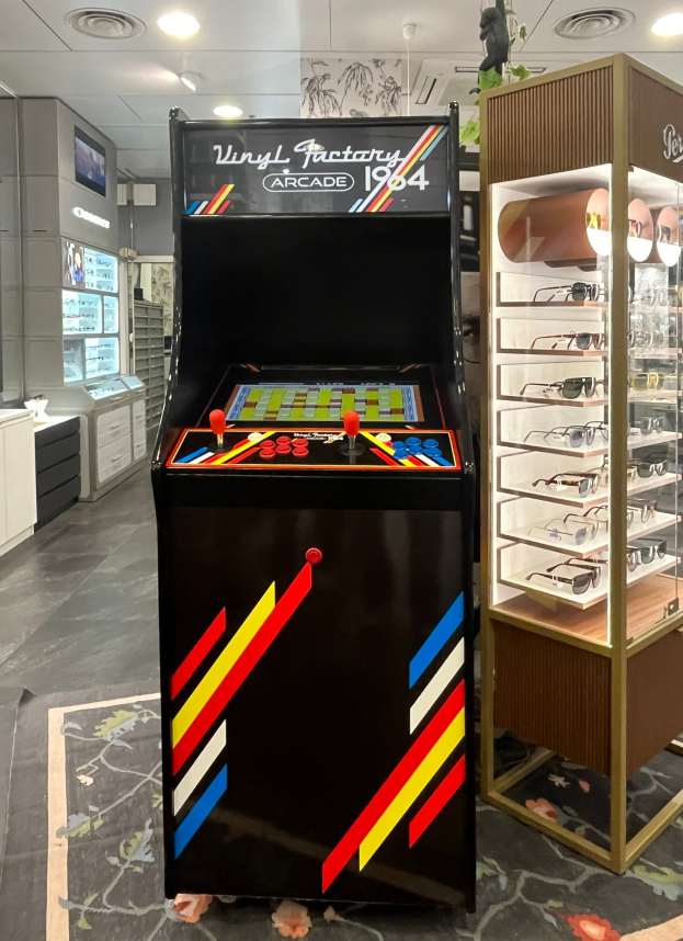 Ein Geschäft mit einem zentralen Vintage-Arcade-Automaten, einem Gestell mit Gläsern auf der rechten Seite und Hintergrund-Elementen wie Schränken, einem Fernseher und anderen Gegenständen, mit Deckenlampen, die eine retro-mäßige Atmosphäre schaffen.
