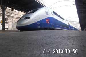 Ein Zug ist in der Mitte des Bildes zu sehen, mit einer Plattform und Text unten, Gebäuden und einem Dach links und Gebäuden, Dächern, Kabeln und Himmel oben.