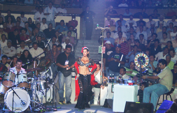 Eine Person spielt ein Musikinstrument auf der B├╝hne, ein Mann in einem schwarzen Hemd hÃ¤lt ein Mikrofon, eine Frau in einem schwarzen und roten Kleid steht neben ihm, eine Person sitzt in einer Ecke und hÃ¤lt eine Kamera, das Publikum sitzt der B├╝hne gegen├╝ber, ein Tisch mit GlÃ¤sern und Flaschen ist vor dem Publikum, und ein Lautsprecher befindet sich unten im Bild.