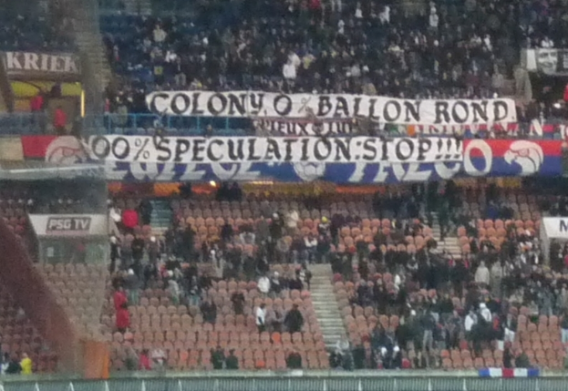 Ein Stadion mit einer gro√üen Menge, Bannern und St√ľhlen, einem Zaun unten und einer Treppe links.