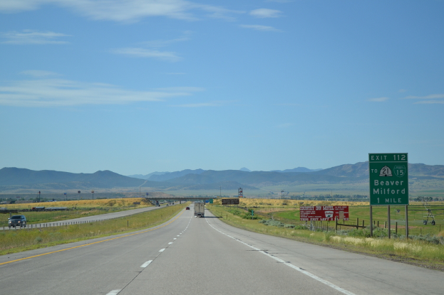 Eine Landschaft mit einem bewölkten Himmel, Hügel, einer Straße mit Kraftfahrzeugen, Barrieren, Schildern, Informationsanzeigen, landwirtschaftlichen Höfen und Grasflächen.