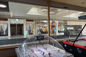 Eine Eishalle in einem Einkaufszentrum, umgeben von Glaswänden mit Menschen, Möbeln und Beschriftung draußen, mit einem Air-Hockey-Tisch auf der rechten Seite.