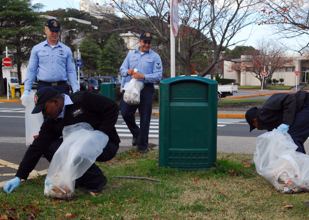 Drei Polizisten in Uniform sammeln Müll neben einer Straße mit Bäumen, Gebäuden und Fahrzeugen im Hintergrund.