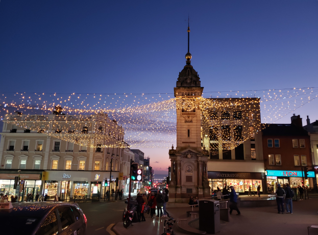 Eine belebte Stadtstraße bei Nacht mit Menschen, Fahrzeugen, Fahrrädern, Mülltonnen, Gebäuden, Laternen, Verkehrsampeln und einem Uhrenturm im Hintergrund, beleuchtet von festlichen Weihnachtslichtern.