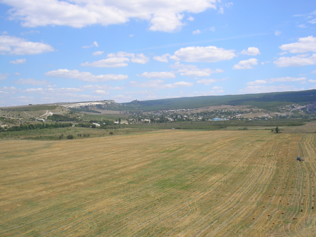 Luftaufnahme eines Traktors bei der Arbeit in einem kultivierten Feld, umgeben von Bäumen, Gebäuden, Hügeln und einer bewölkten Himmelslandschaft.