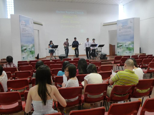 Eine Bühne mit zwei Gitarristen in der Mitte und anderen, die singen und Instrumente spielen, in den Ecken, mit Bannern an den Seiten und einem Publikum davor, das Papiere und Bücher hält.