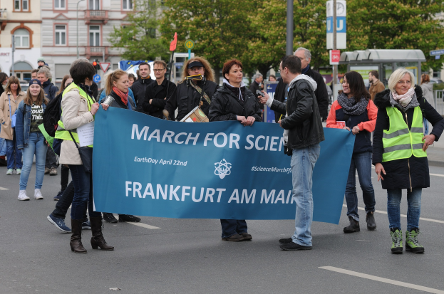 Eine bunte Gruppe von Menschen marschiert eine Straße entlang, hält ein "Marsch für die Wissenschaft Frankfurt am Main"-Schild hoch, mit Bäumen, Pfählen, Schildern, Gebäuden und einem klaren blauen Himmel im Hintergrund.