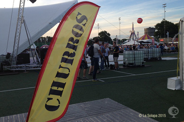 Ein buntes Karnevalsszenario mit Zelten, einem zentralen Mast, Menschen, Bäumen, Strommasten, Straßenbahnen auf der Straße und einem eisernen Stand auf der linken Seite unter einem klaren Himmel.