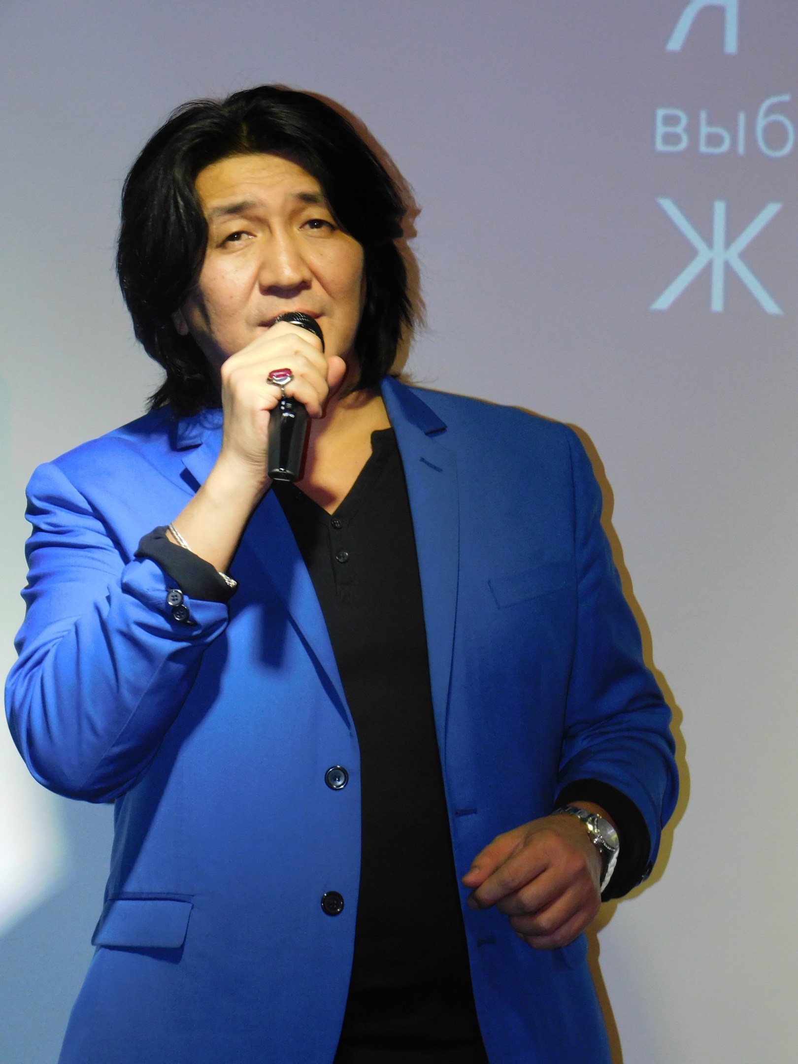 Ein Mann in einem blauen Anzug hält ein Mikrofon und steht vor einem Bildschirm mit Text, der wie ein Regisseur mit ernster Miene aussieht.