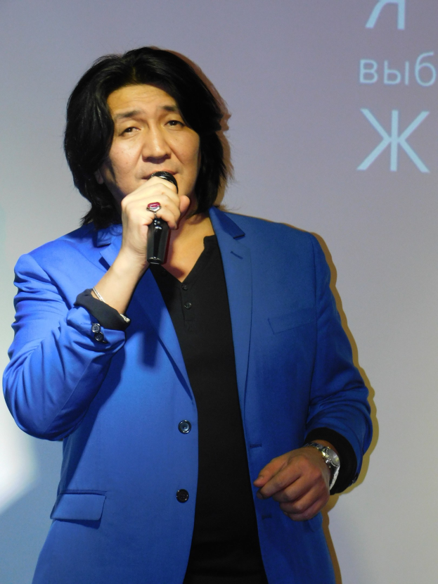 Ein Mann in einem blauen Anzug hält ein Mikrofon und steht vor einem Bildschirm mit Text, der wie ein Regisseur mit ernster Miene aussieht.