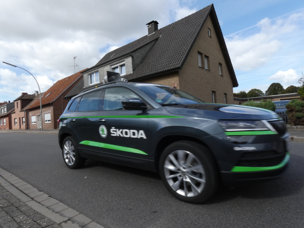 Schwarzer und grüner Skoda Kodiaq Elektroauto fährt auf einer Straße mit Gebäuden, Laternen, Bäumen und einem bewölkten Himmel im Hintergrund.