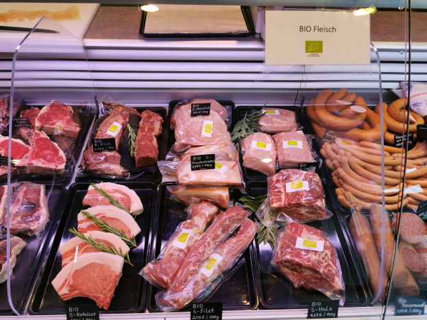Vitrine mit verschiedenen Fleisch- und Wurstsorten in Schalen mit Preisschildern in einer Metzgerei mit einem Schild im Hintergrund.