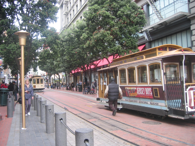 Eisenbahnschienen verlaufen entlang einer Straße mit darauf fahrenden Zügen, einer davon mit Text, und einer Person in der Nähe; kleine Pfosten mit Seilen und Straßenlaternen säumen die linke Seite, während Bäume, viele Menschen, ein Müllcontainer und Gebäude mit Leitern auf der rechten Seite zu sehen sind.