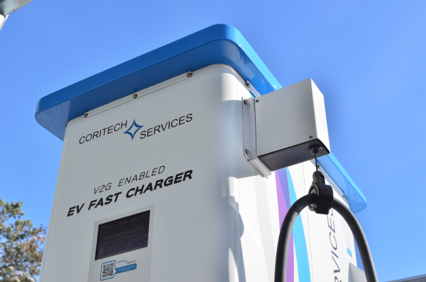 Elektrofahrzeug-Ladestation mit der Beschriftung "V2G Fast Charger" im Freien mit B├Ąumen und einem Geb├Ąude im Hintergrund bei klarem blauem Himmel.
