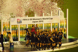 Eine Gruppe von Frauenfußballspielerinnen in schwarzen Uniformen steht auf einer Bühne und feiert ihren Sieg, umgeben von einer Menge mit Kameras, mit einem dekorierten Hintergrund und grasbewachsenem Boden.