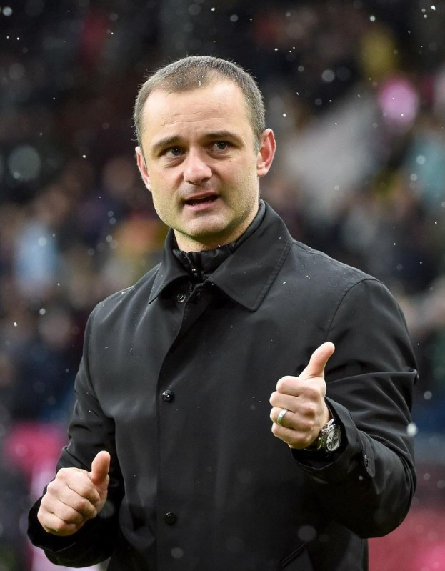 Ein Mann in einer schwarzen Jacke zeigt den Daumen hoch, trägt eine Uhr, mit einem unscharfen Hintergrund.