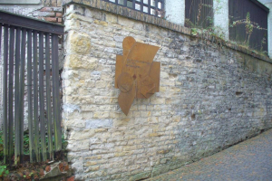 Steinmauer mit Metalltor auf der linken Seite, eine Gedenktafel für die Einweihung des alten Rathauses und ein Gebäude mit Fenstern und Pflanzen im Hintergrund.