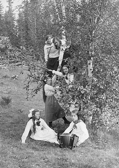 Schwarzes und weißes Foto von jungen Mädchen, die Akkordeons im Freien in einer grasbewachsenen Fläche mit Bäumen und einem Zaun im Hintergrund spielen.