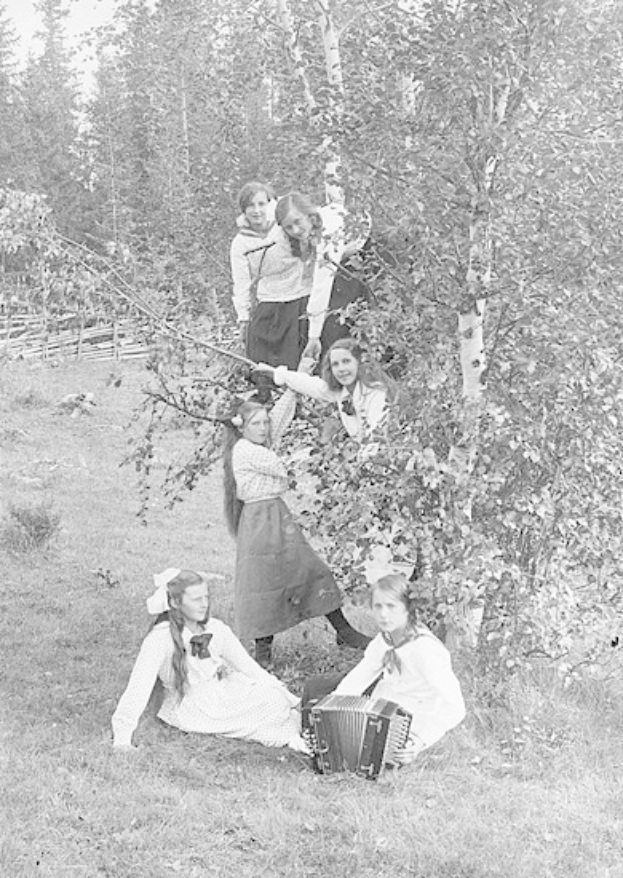 Schwarzes und weißes Foto von jungen Mädchen, die Akkordeons im Freien in einer grasbewachsenen Fläche mit Bäumen und einem Zaun im Hintergrund spielen.