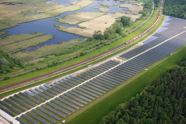 Luftaufnahme einer Solaränlage mit Panels, umgeben von Bäumen, Gras, Pflanzen und Wasser, mit einem Zug auf einer näheren Bahnstrecke.