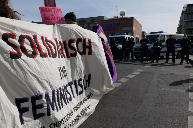 Eine Gruppe von Menschen marschiert auf einer Straße, hält ein Banner mit der Aufschrift "Solidarität und Feminismus" in der Hand, mit parkenden Fahrzeugen und Gebäuden im Hintergrund unter einem klaren blauen Himmel.