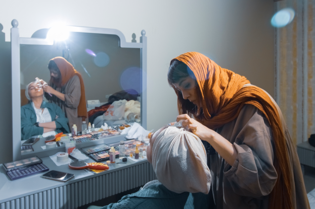 Eine Frau sitzt vor einem Schminktisch mit Spiegel auf dem Boden, trägt ein Kopftuch und hält ein Objekt in der Hand, umgeben von Make-up-Bürsten, Flaschen, einem Handy, einer Rechenmaschine und anderen Gegenständen, mit einer Lampe an der Wand und einem Vorhang auf der rechten Seite.
