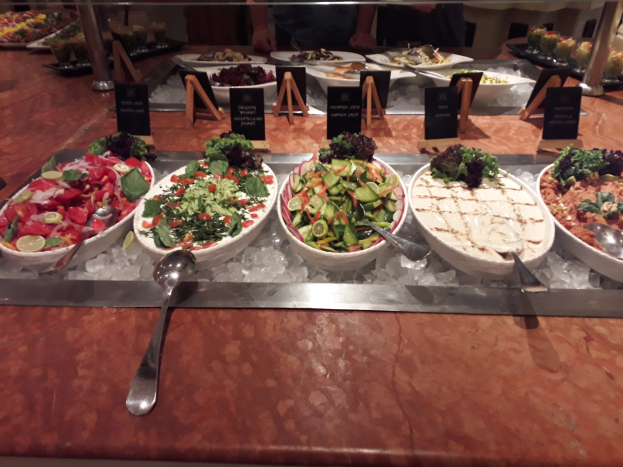 Ein Buffet mit verschiedenen Gerichten, darunter Salate, Bestecke und Eiswürfel, begleitet von kleinen Informationsschildern und Menschen im Hintergrund.